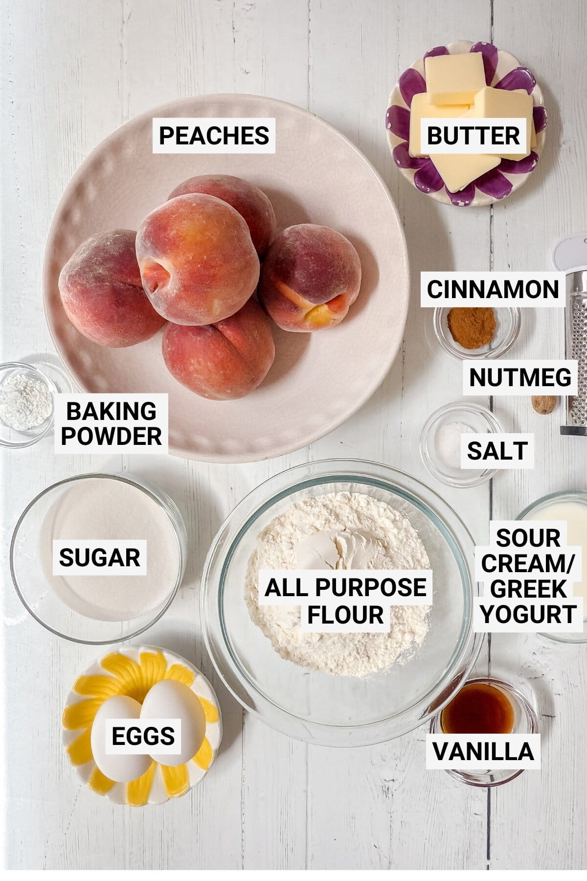 Top-down view of labeled baking ingredients for peach cobbler cake on a white table, including peaches, butter, cinnamon, nutmeg, salt, baking powder, sugar, eggs, all-purpose flour, sour cream or Greek yogurt, and vanilla.