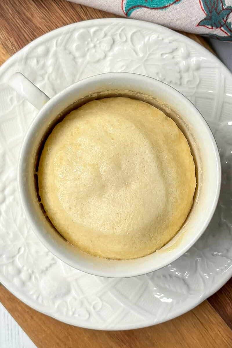 A white cup filled with Pineapple Upside Down Mug Cake sits on a matching patterned saucer atop a wooden board, with a colorful floral cloth in the background.