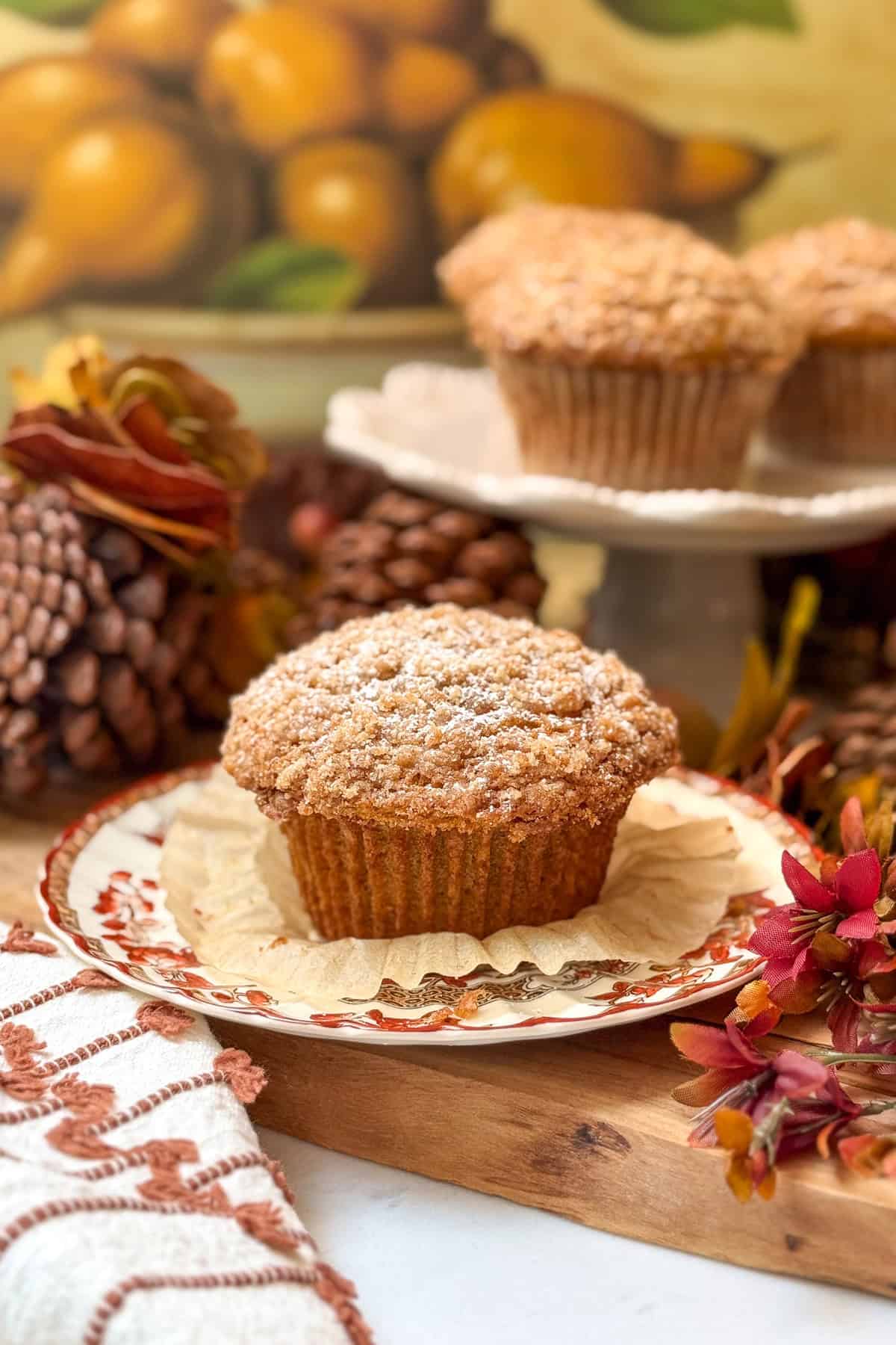 A single Panera pumpkin muffin with a crumb topping sits on a decorative plate, with another muffin on a stand in the background.