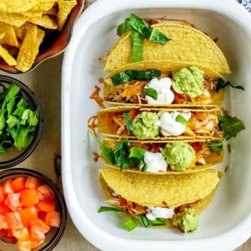 Three hard-shell tacos in a dish, topped with lettuce, cheese, salsa, sour cream, and guacamole—a perfect addition to your Back to School Dinner Recipes.