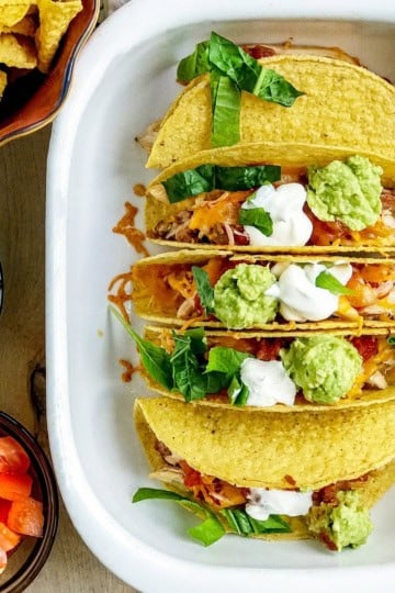 Three hard-shell tacos in a dish, topped with lettuce, cheese, salsa, sour cream, and guacamole—a perfect addition to your Back to School Dinner Recipes.