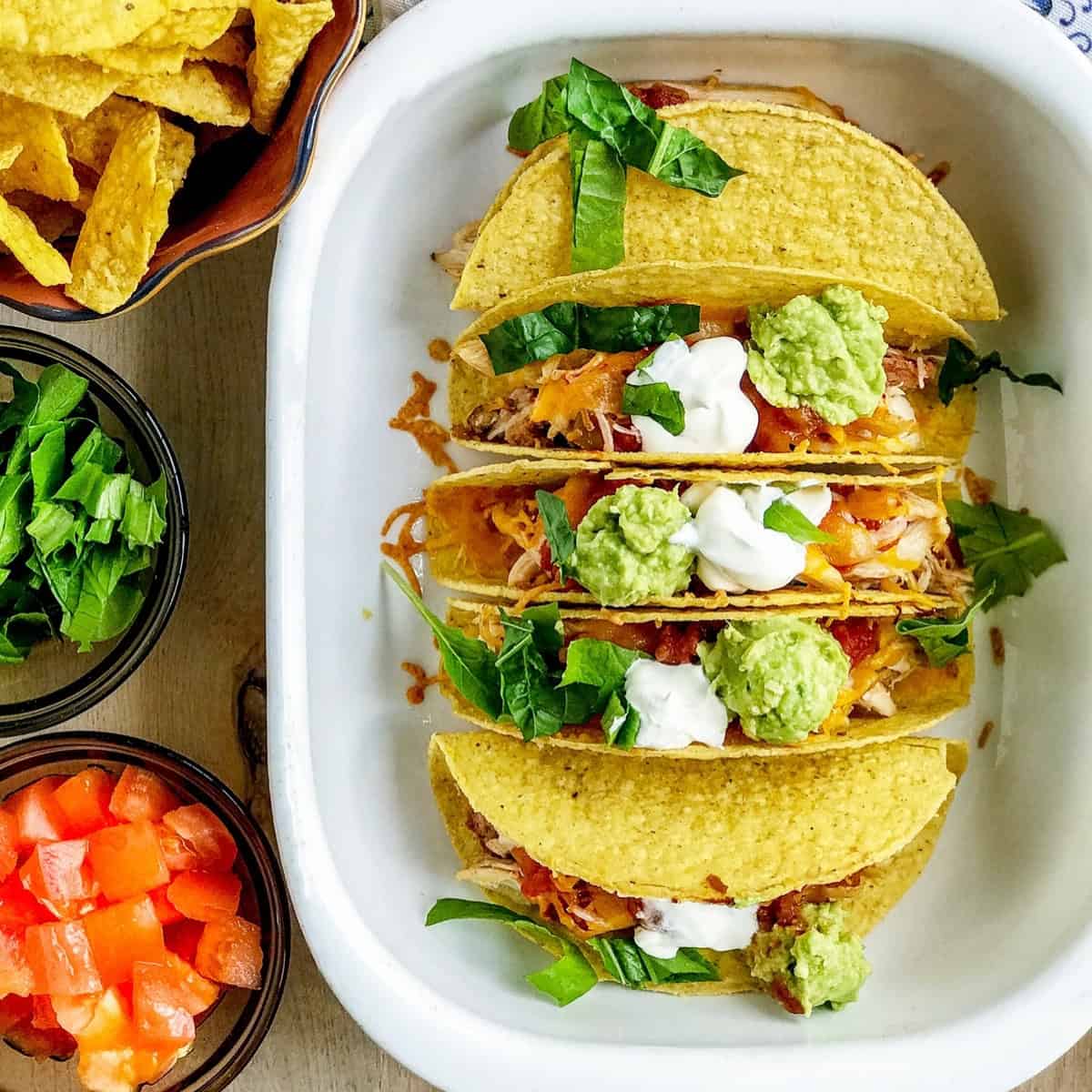 Three hard-shell tacos in a dish, topped with lettuce, cheese, salsa, sour cream, and guacamole-a perfect addition to your Back to School Dinner Recipes.
