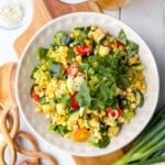 A bowl of mixed vegetable salad with grilled corn, zucchini, tomatoes, cucumbers, greens, and cilantro on a white plate, set on a wooden board with utensils and green onions nearby.