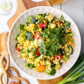 A bowl of mixed vegetable salad with grilled corn, zucchini, tomatoes, cucumbers, greens, and cilantro on a white plate, set on a wooden board with utensils and green onions nearby.