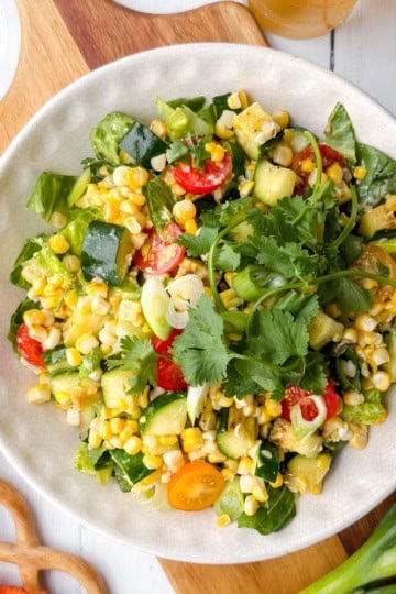A bowl of mixed vegetable salad with grilled corn, zucchini, tomatoes, cucumbers, greens, and cilantro on a white plate, set on a wooden board with utensils and green onions nearby.