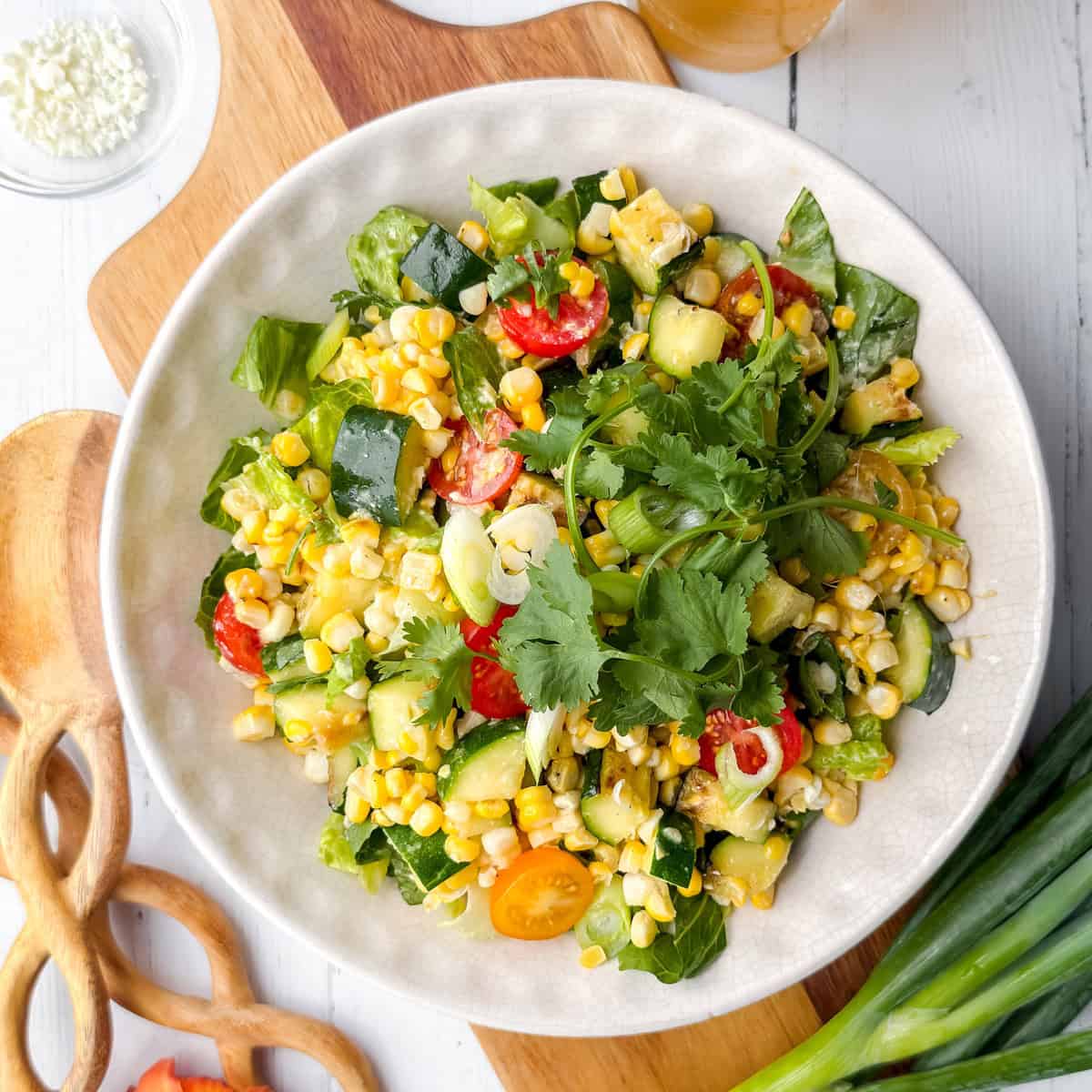 A bowl of mixed vegetable salad with grilled corn, zucchini, tomatoes, cucumbers, greens, and cilantro on a white plate, set on a wooden board with utensils and green onions nearby.