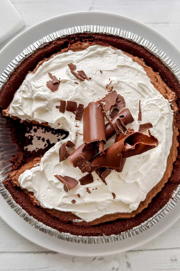 A no bake chocolate pie in a foil tin with a slice missing, this decadent chocolate pie is topped with whipped cream and chocolate shavings, served on a white plate.