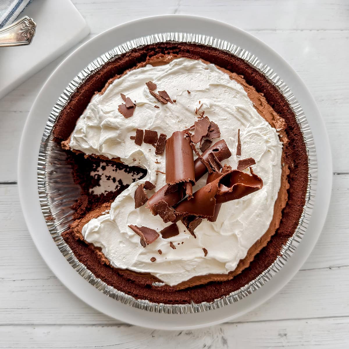 A no bake chocolate pie in a foil tin with a slice missing, this decadent chocolate pie is topped with whipped cream and chocolate shavings, served on a white plate.