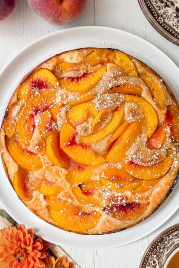 A round peach cobbler cake topped with powdered sugar sits on a white plate, surrounded by peaches, a striped towel, and a cup of tea on a white wooden table.