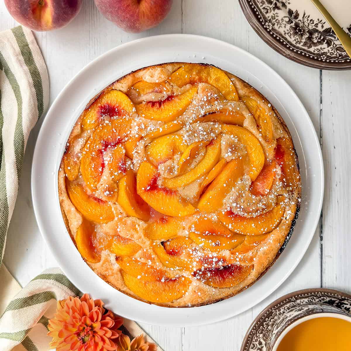 A round peach cobbler cake topped with powdered sugar sits on a white plate, surrounded by peaches, a striped towel, and a cup of tea on a white wooden table.