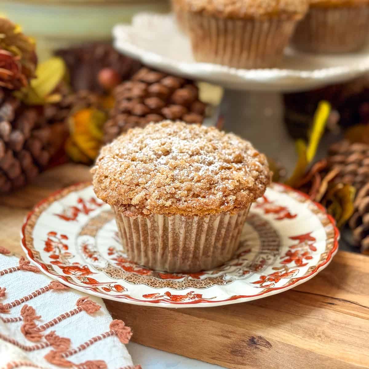 A Pumpkin Muffin topped with powdered sugar sits on a decorative plate, reminiscent of Panera, with pinecones and more muffins in the background.