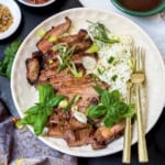 A plate with sliced grilled steak and rice, garnished with herbs, green onions, and fresh basil, served with a fork and knife.