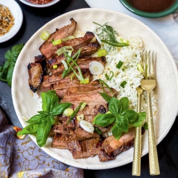 A plate with sliced grilled steak and rice, garnished with herbs, green onions, and fresh basil, served with a fork and knife.