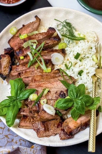 A plate with sliced grilled steak and rice, garnished with herbs, green onions, and fresh basil, served with a fork and knife.