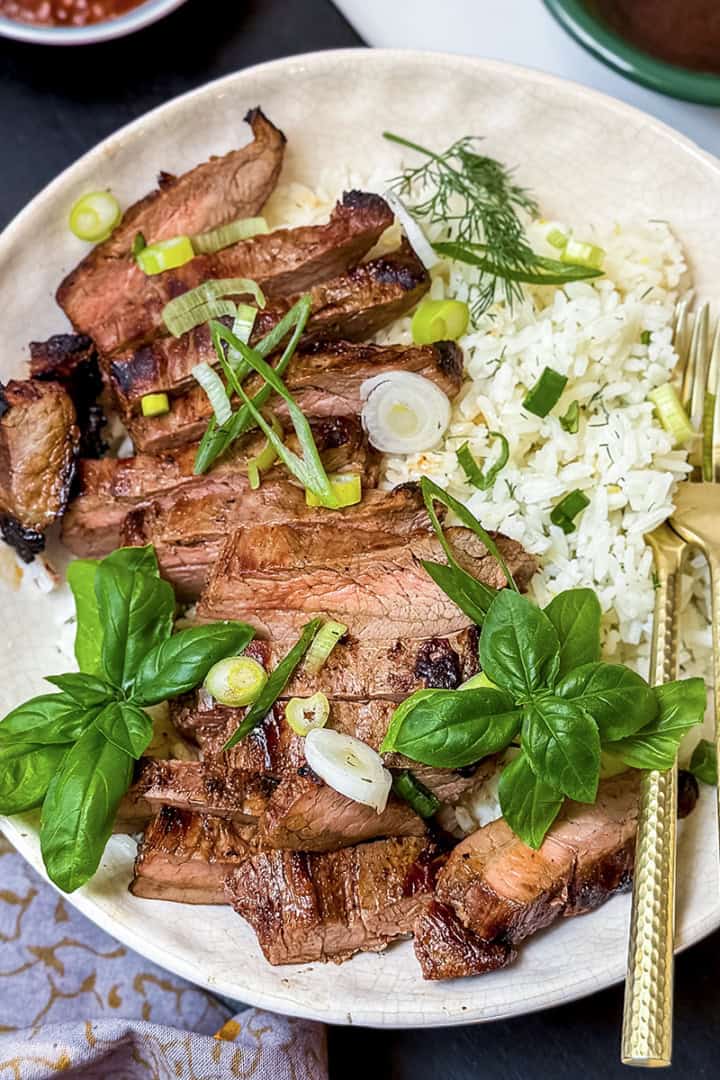 A plate with sliced grilled steak and rice, garnished with herbs, green onions, and fresh basil, served with a fork and knife.