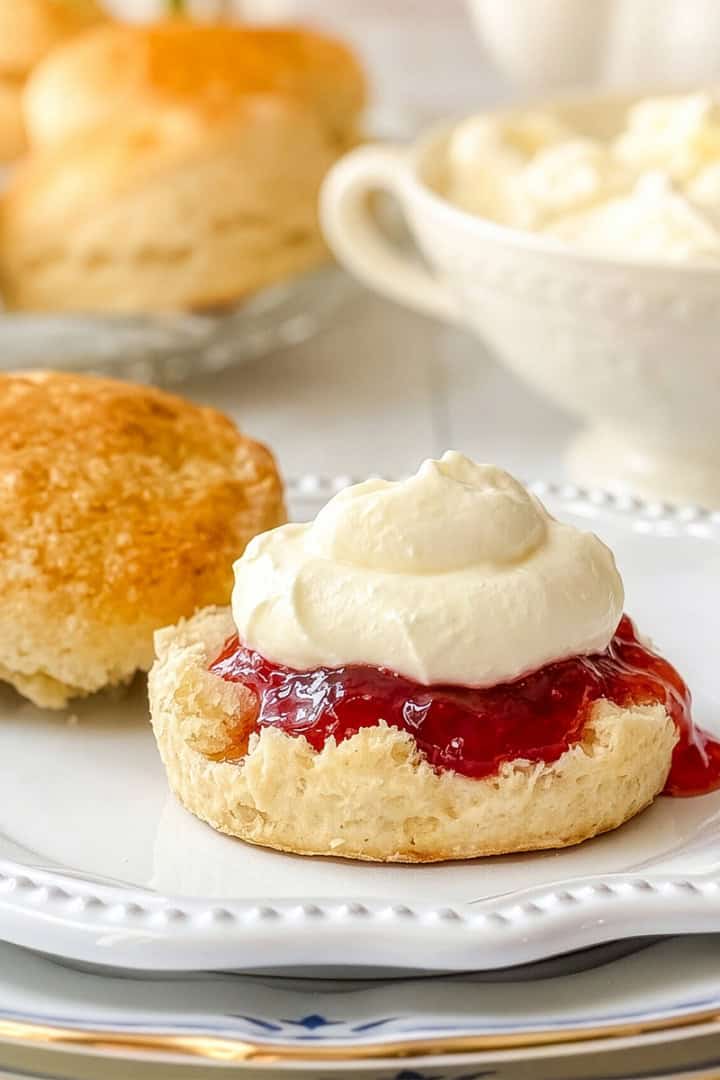A scone cut in half on a plate, with one half topped with red jam and a dollop of homemade clotted cream, and the other half plain; more scones and a bowl of cream are in the background.