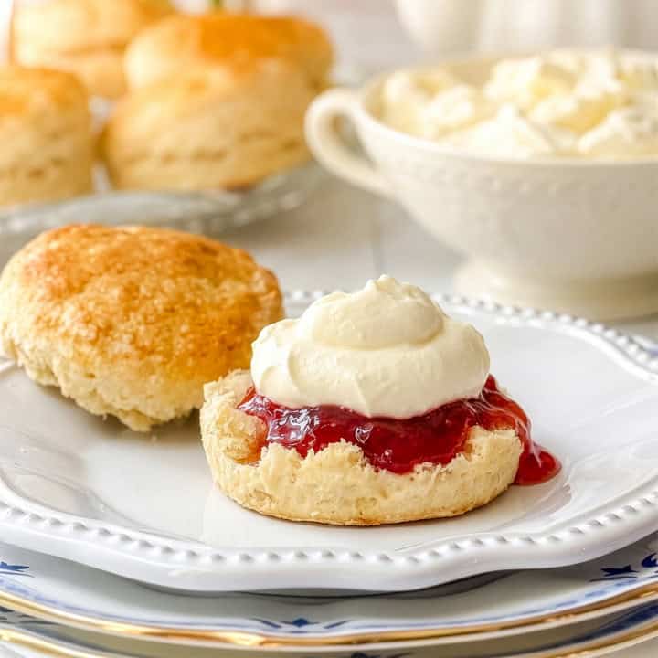 A scone cut in half on a plate, with one half topped with red jam and a dollop of homemade clotted cream, and the other half plain; more scones and a bowl of cream are in the background.