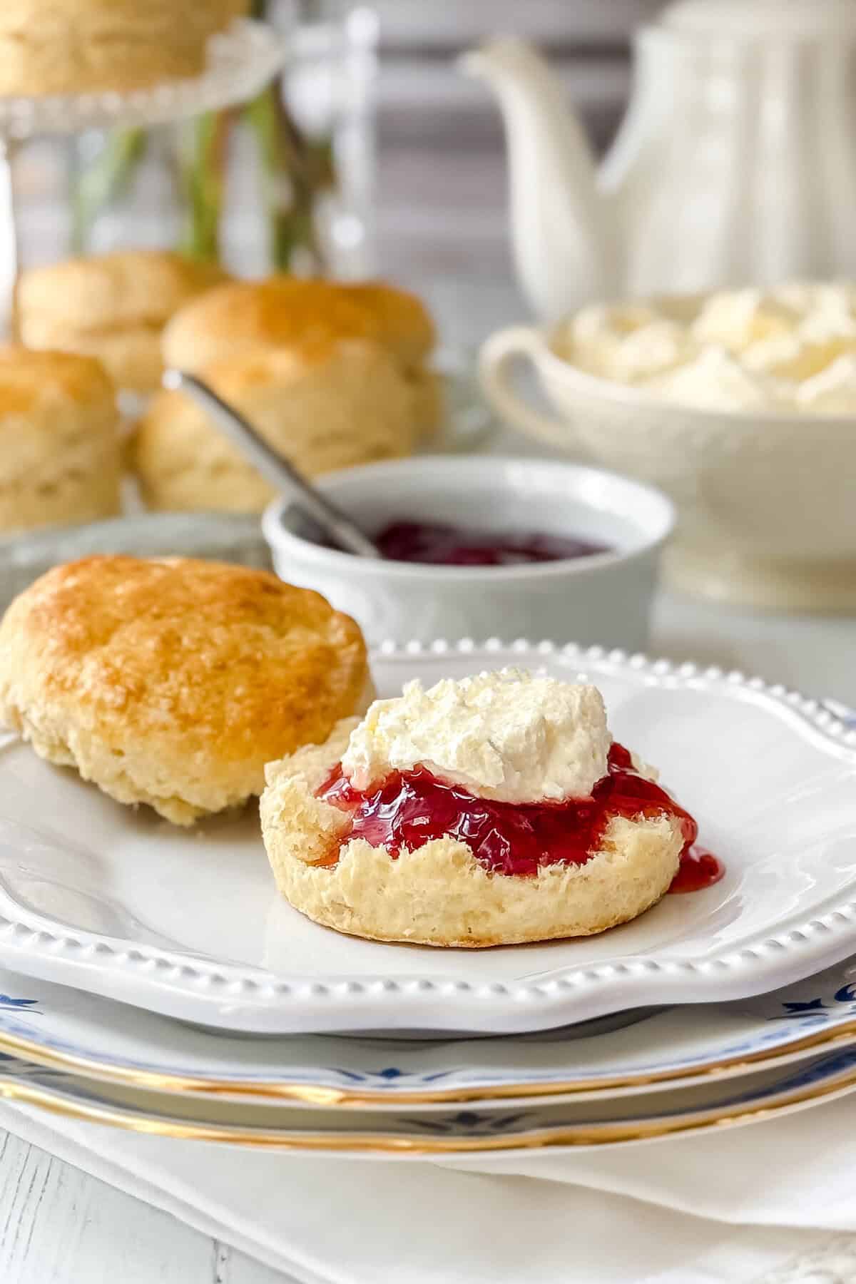 A scone topped with homemade clotted cream and strawberry jam sits on a white plate, surrounded by more scones, a bowl of jam, clotted cream, and a teapot—the perfect treat for fans of 3 ingredient recipes.