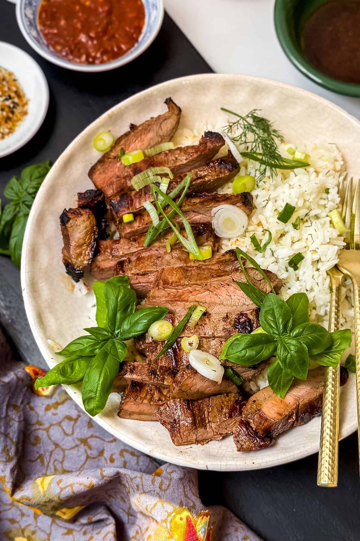 A plate of sliced Lemon Ginger Teriyaki Steak served over white rice, garnished with fresh basil and green onions, with small bowls of sauce and seasonings on the side-perfect for quick 30 Minute Recipes.