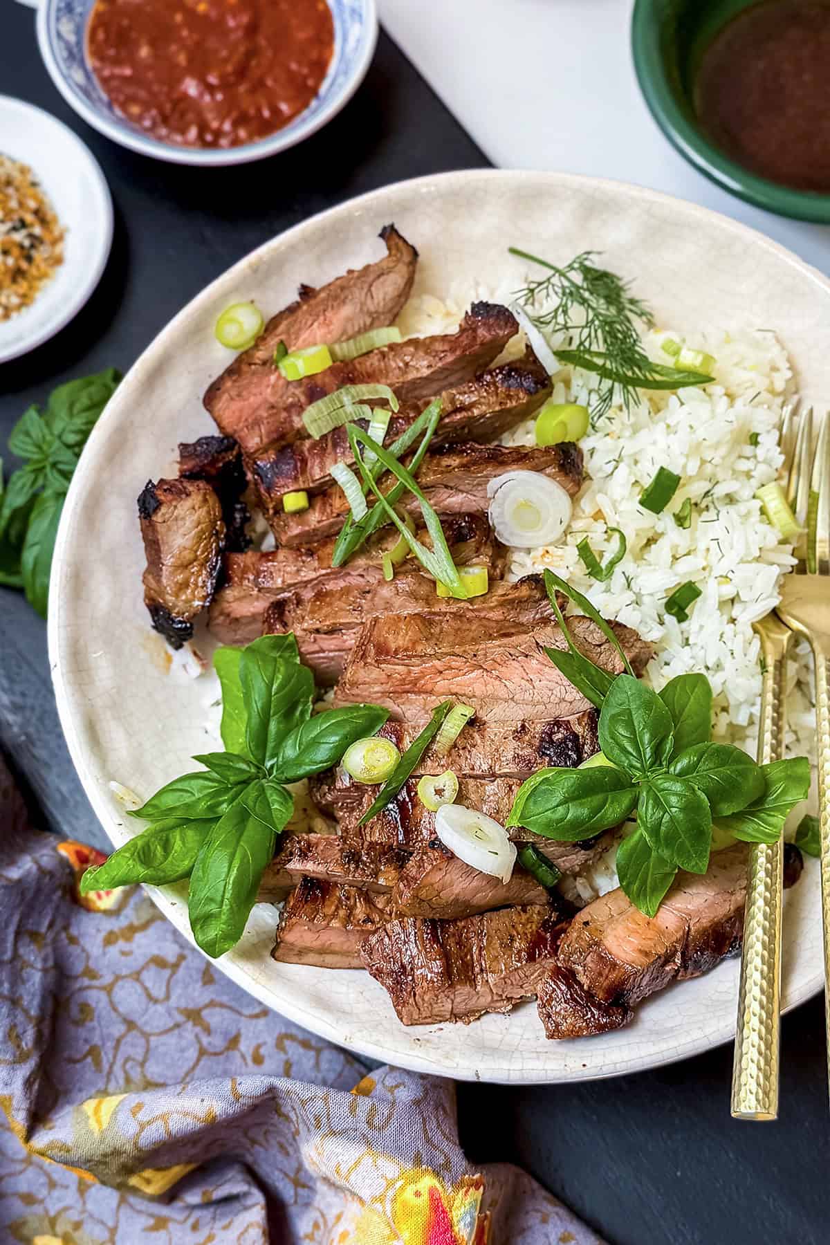 Sliced grilled steak and rice served on a plate with fresh basil, green onions, a fork, and small bowls of sauce and seasonings for the perfect flavor pairing.