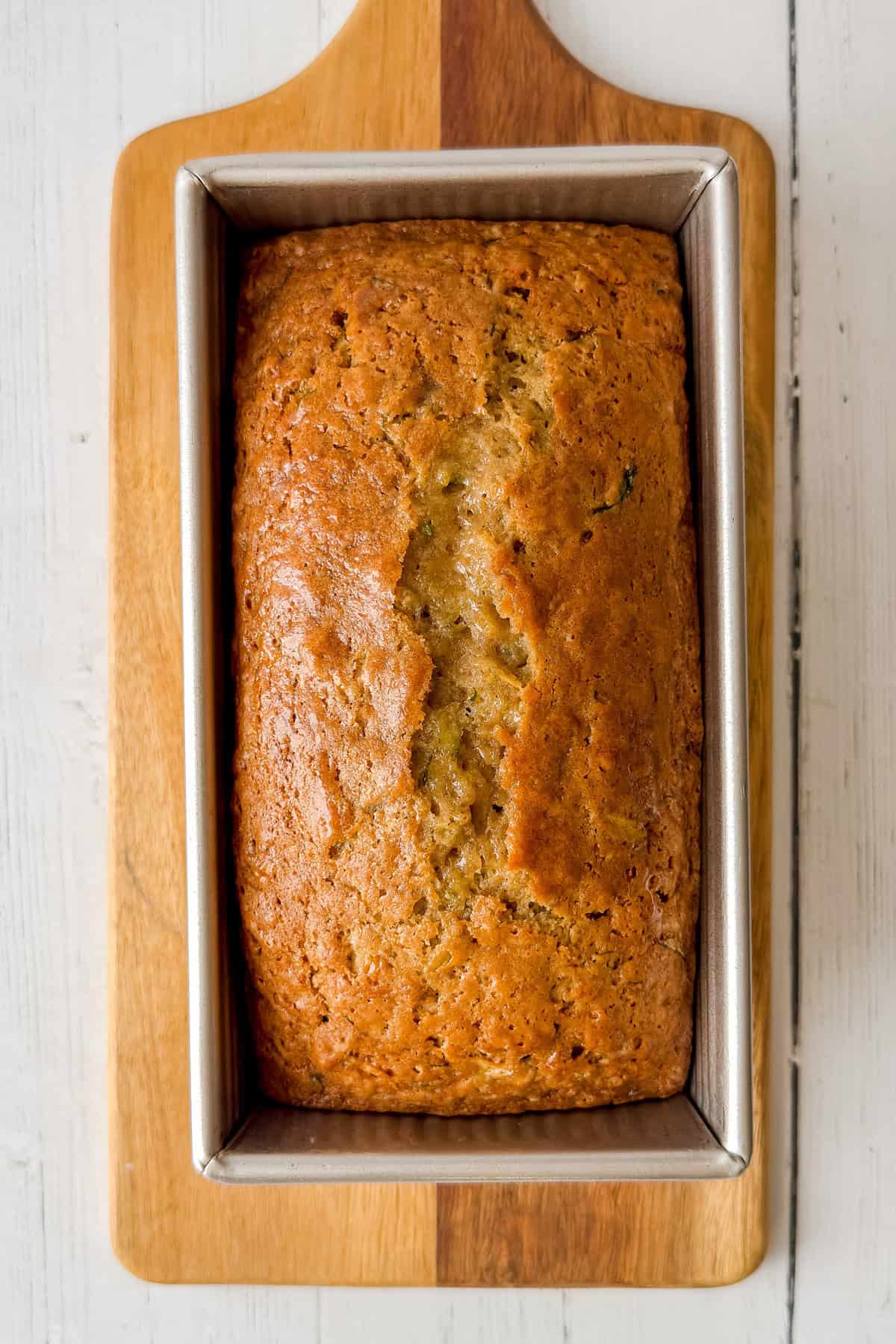 Golden brown single loaf zucchini bread cooling in an 8x4 loaf pan on a wooden cutting board.