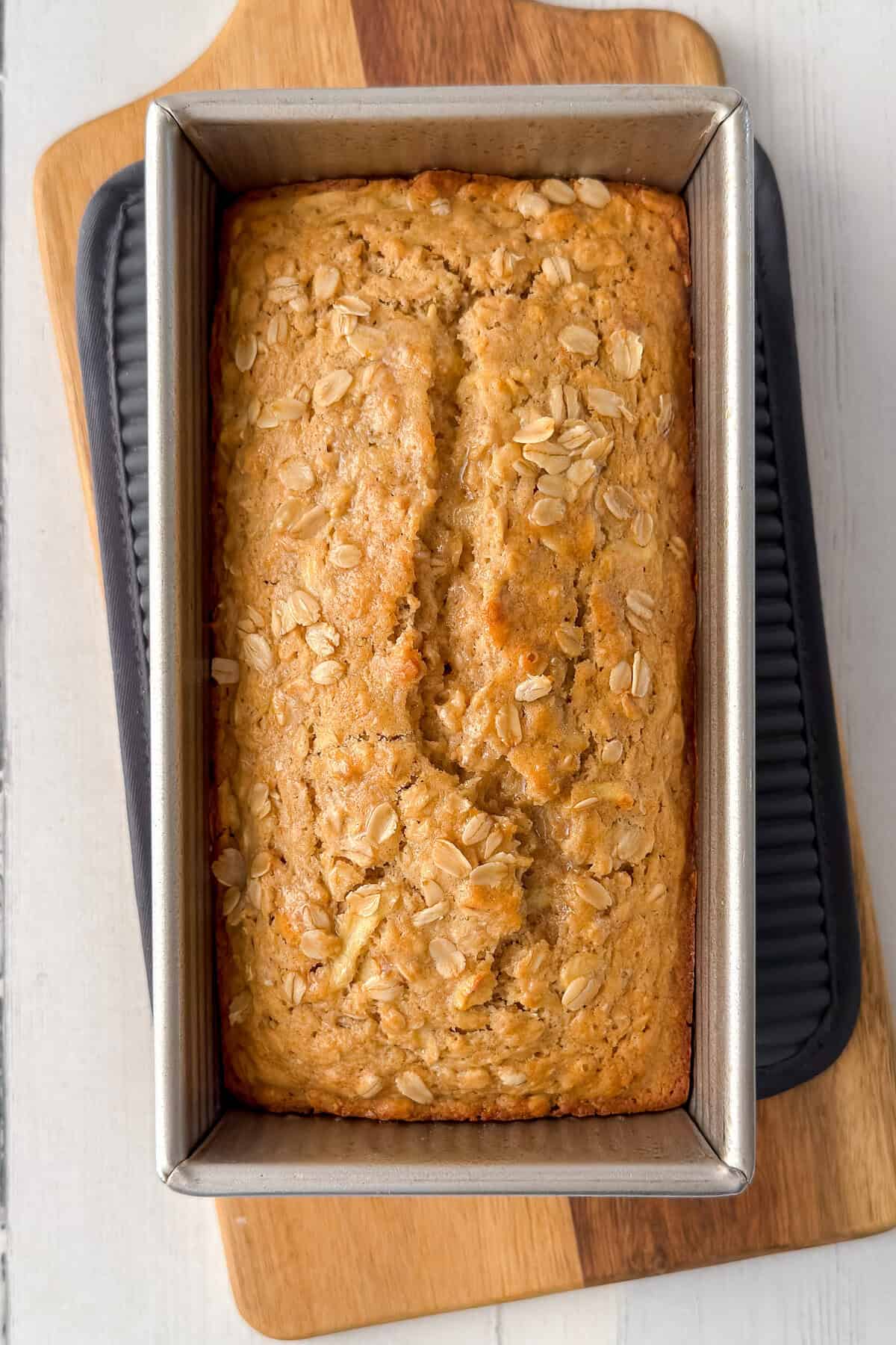 A loaf of apple oatmeal bread in a metal baking pan rests on a wooden cutting board.