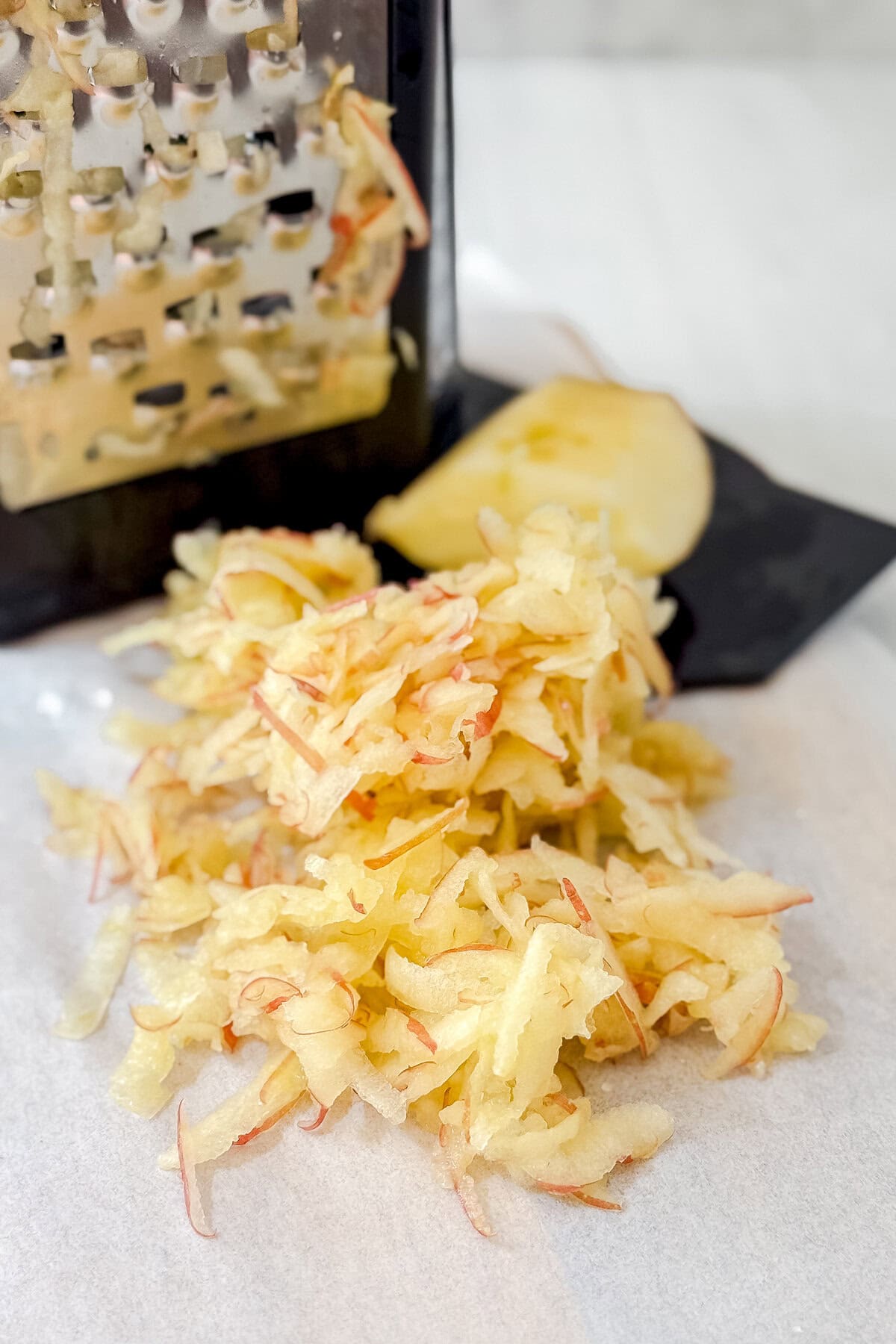 A pile of freshly grated apple on parchment paper, perfect for making apple oatmeal bread, with a box grater and a partially grated apple in the background.