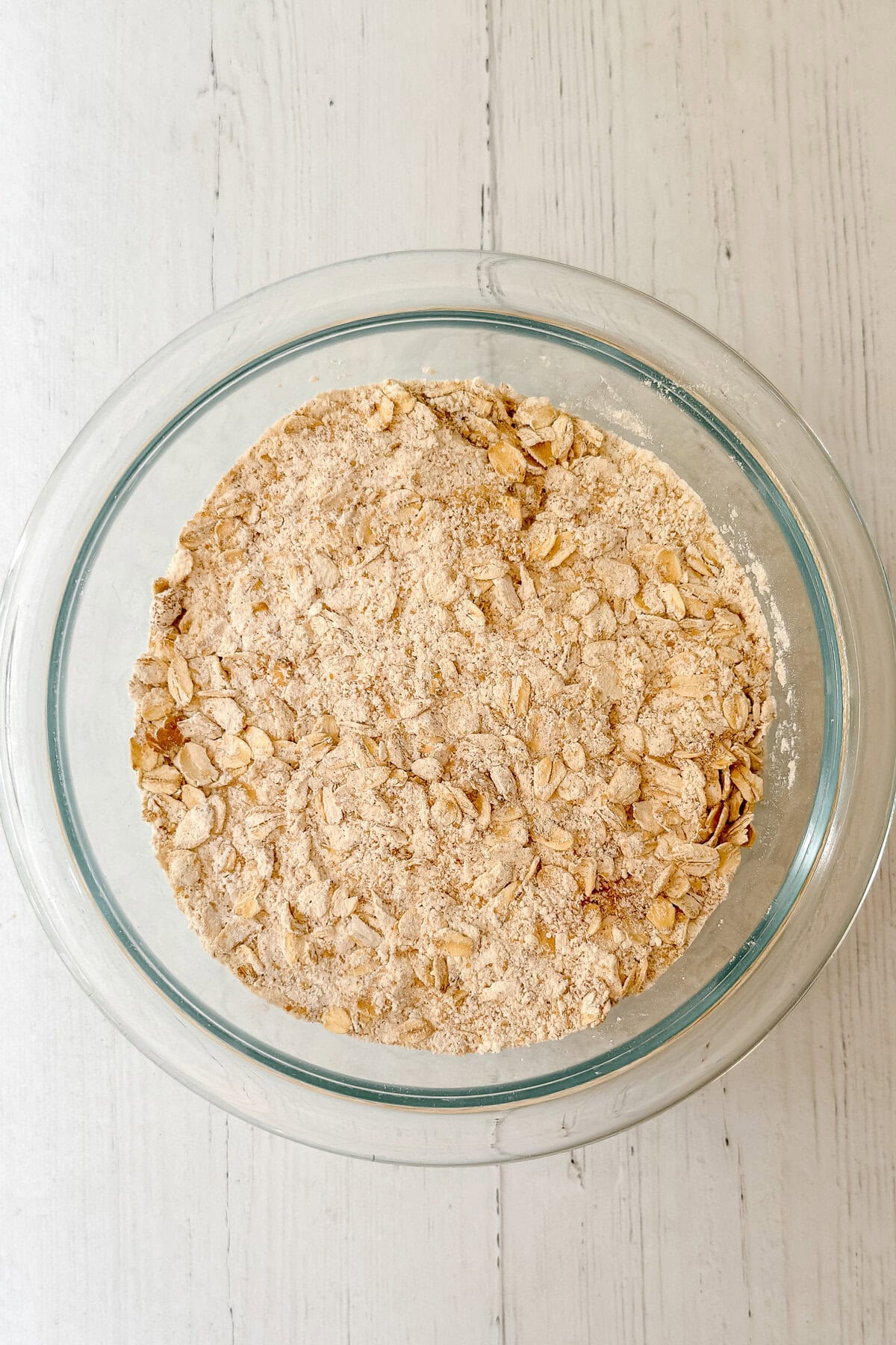 A glass bowl filled with a mixture of oats and flour, ready to become delicious apple oatmeal bread, sits on a white wooden surface.