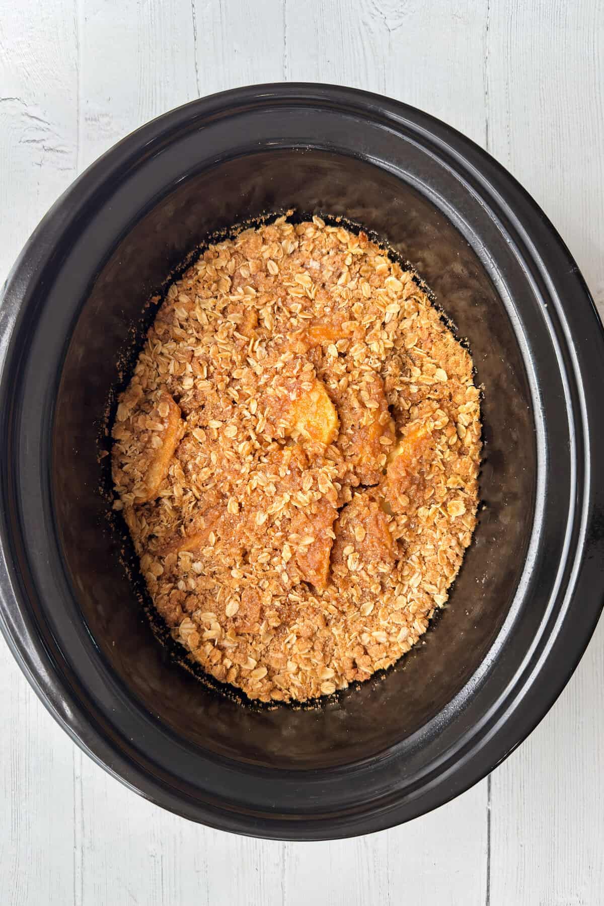 A slow cooker filled with peach and apple crisp, topped with a golden, crumbly oat topping, placed on a white wooden surface.