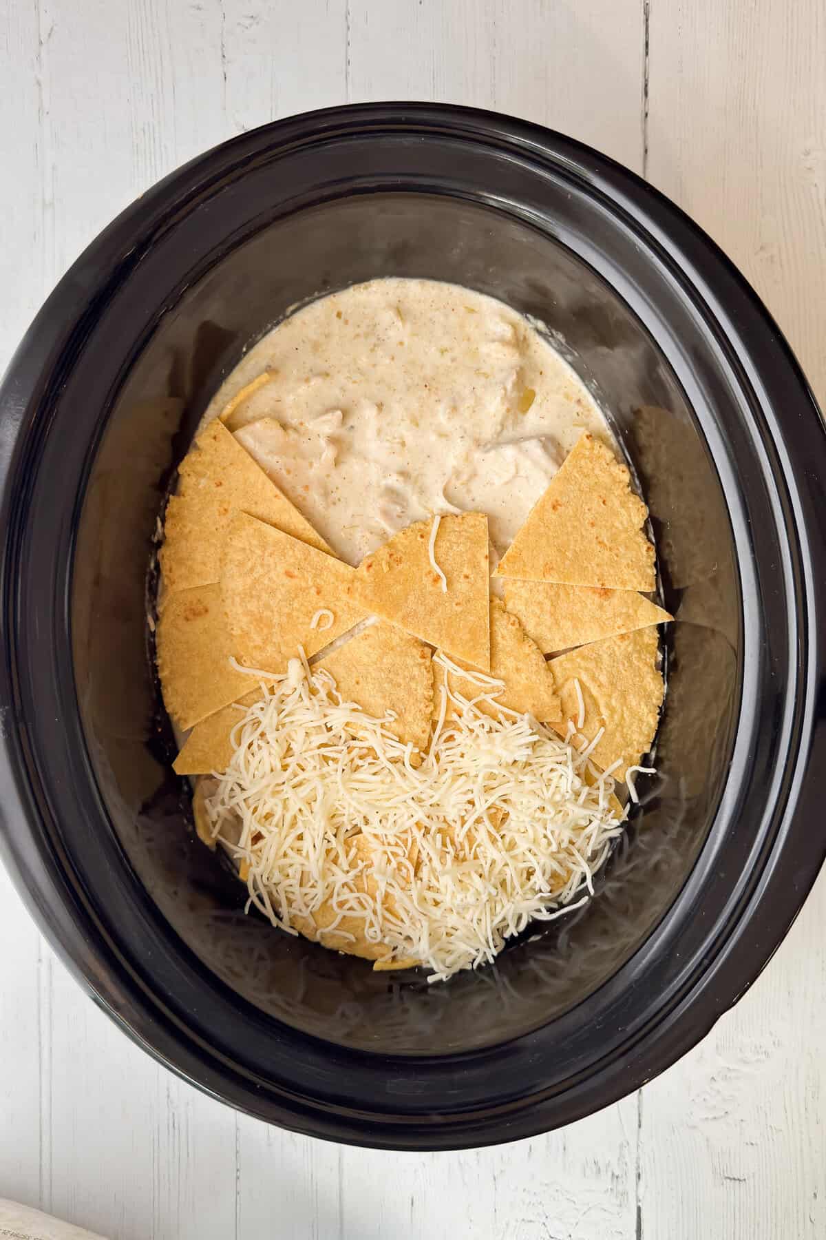 A Crock Pot Chicken Enchilada Casserole with tortilla chips, shredded cheese, and creamy sauce layered inside, sits on a white wooden surface.