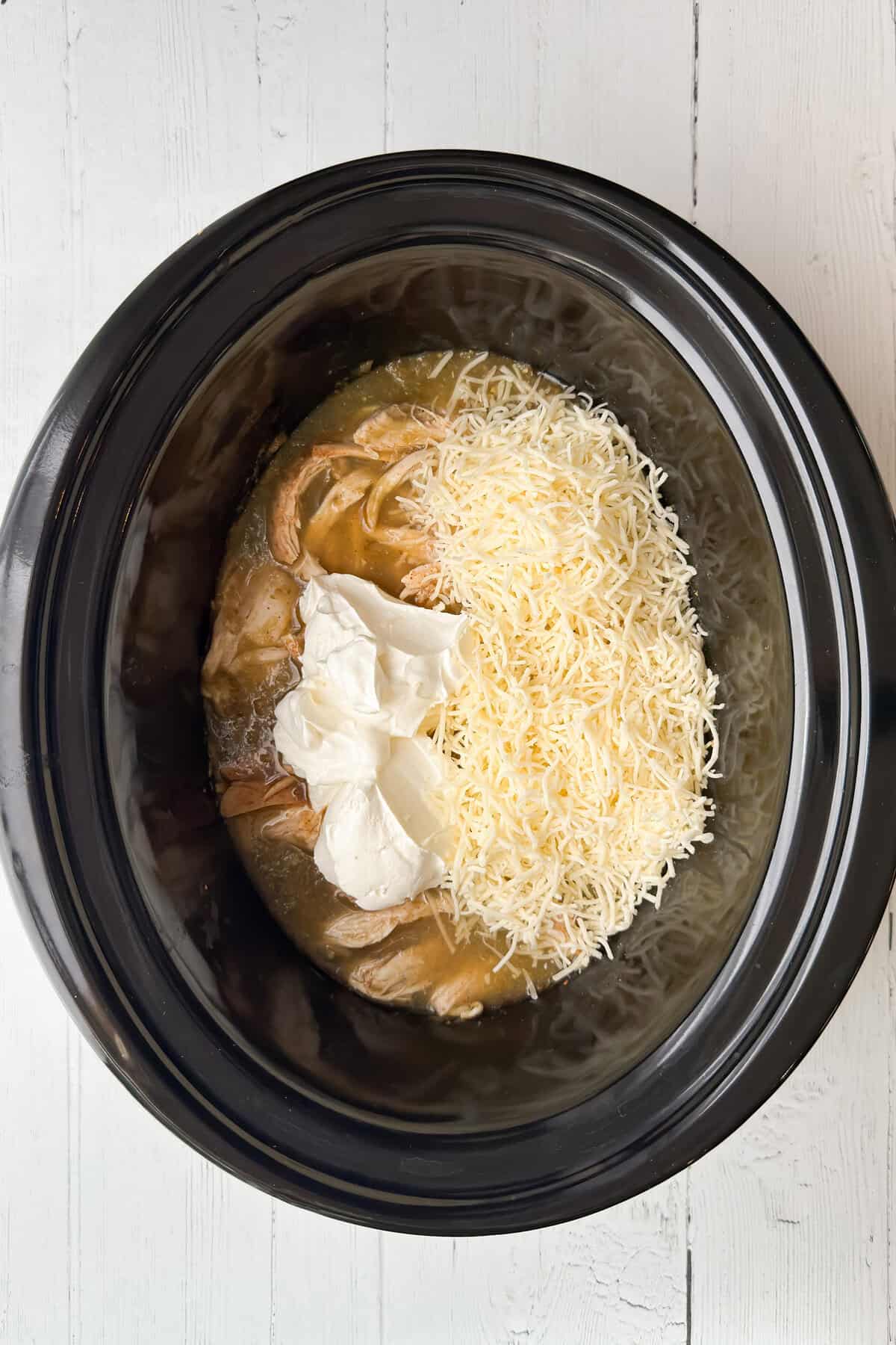 A Crock Pot filled with shredded chicken, shredded cheese, and two scoops of cream cheese creates the perfect base for a Chicken Enchilada casserole on a white wooden surface.