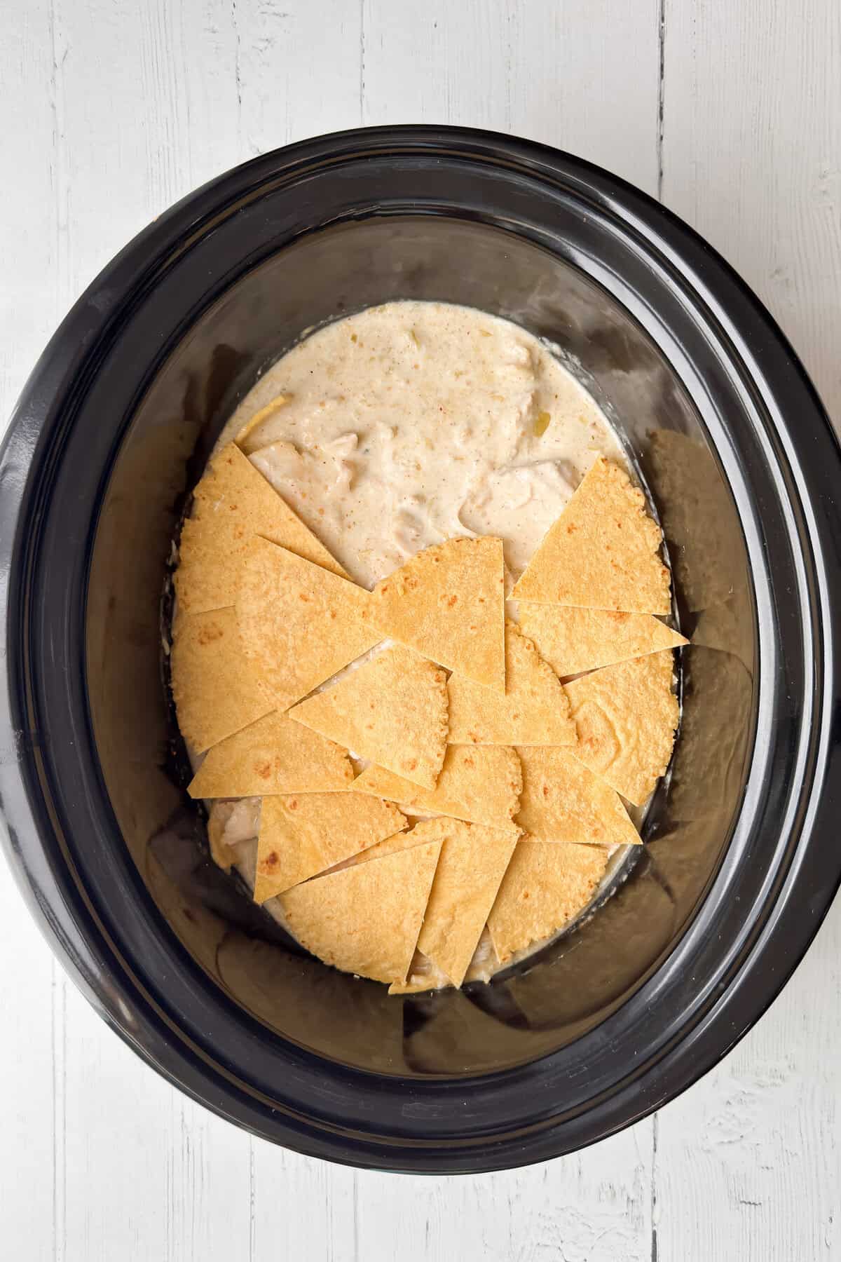 A Crock Pot sits on a white wooden surface, filled with a creamy Chicken Enchilada casserole mixture and layers of corn tortilla pieces.