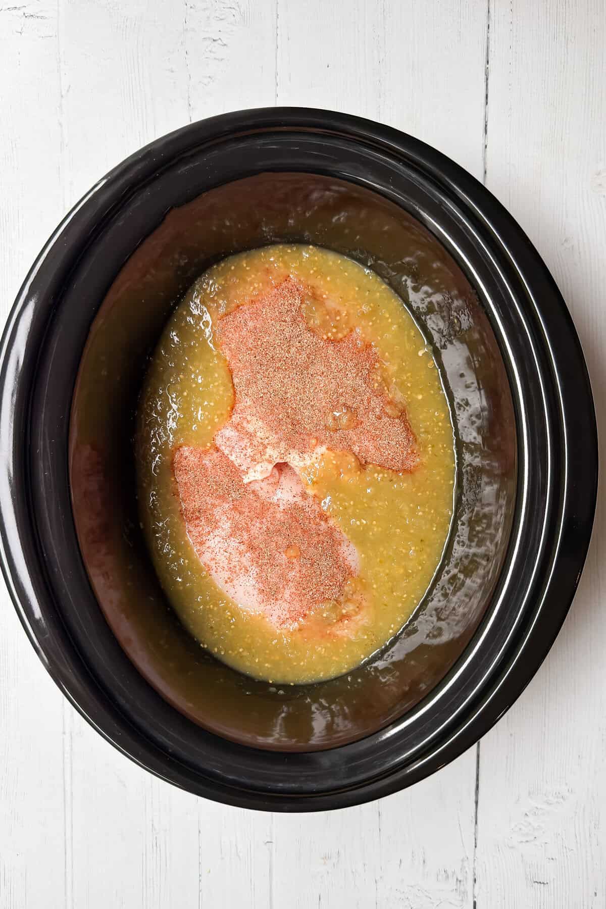 Raw meat with seasoning sits in a Crock Pot, partially covered by a yellowish liquid, on a white wooden surface-perfect for starting a flavorful chicken enchilada casserole.