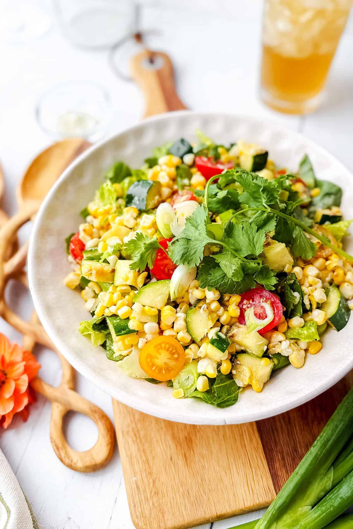 A bowl of fresh Corn Salad with cherry tomatoes, cucumber, greens, cilantro, and grilled zucchini sits on a wooden board next to utensils and a glass of iced tea.