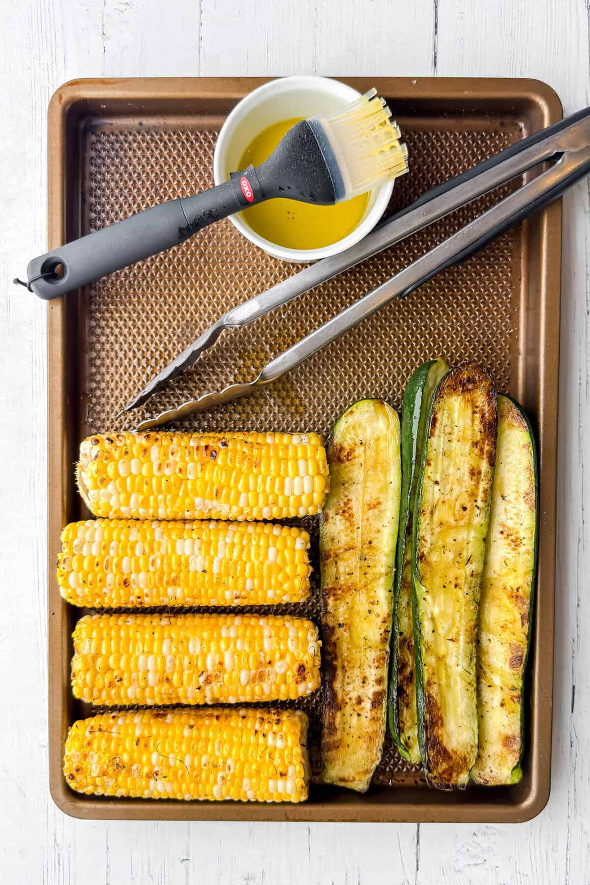 Grilled zucchini and corn on the cob are arranged on a baking sheet with metal tongs, a brush, and a bowl of oil-perfect for a fresh Grilled Zucchini Salad or Corn Salad.