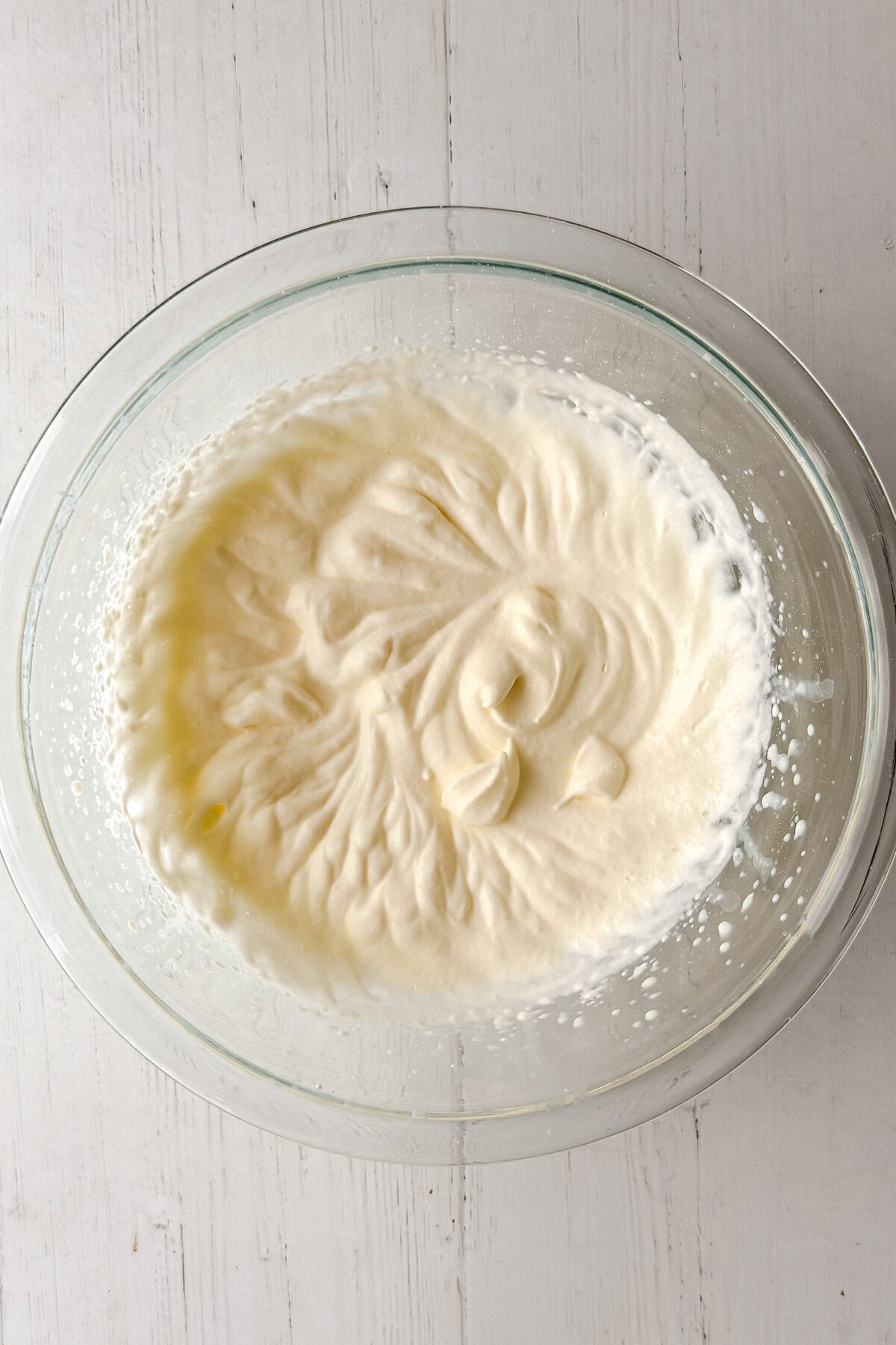 A glass bowl of freshly whipped, homemade clotted cream with soft peaks sits on a white wooden surface, capturing the essence of a classic clotted cream recipe.
