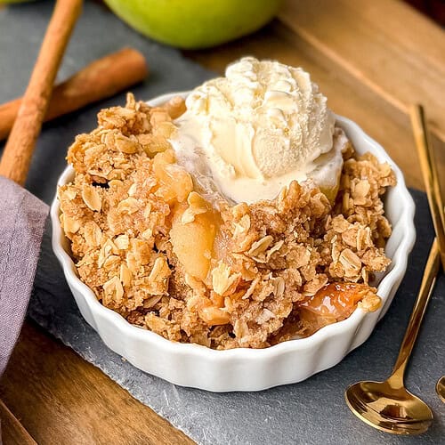 A bowl of slow cooker apple crisp topped with vanilla ice cream, placed on a slate board with two gold spoons and cinnamon sticks nearby.