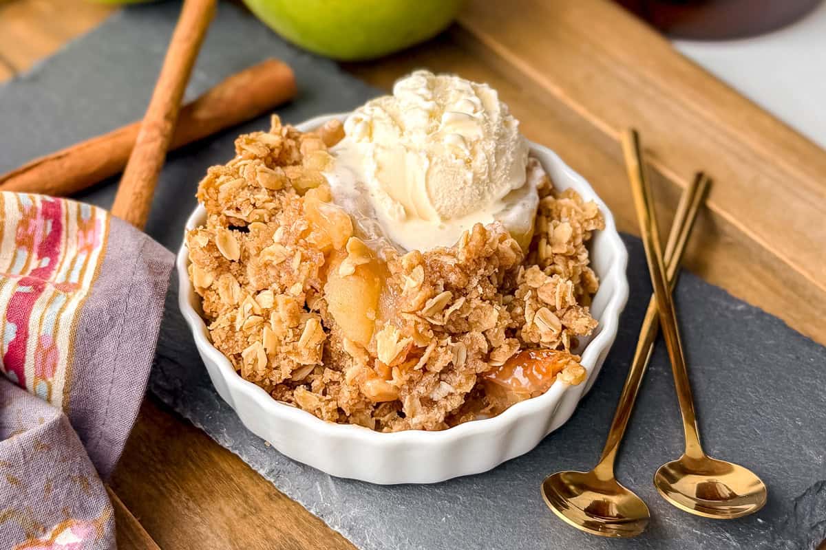 A bowl of slow cooker apple crisp topped with vanilla ice cream, placed on a slate board with two gold spoons and cinnamon sticks nearby.