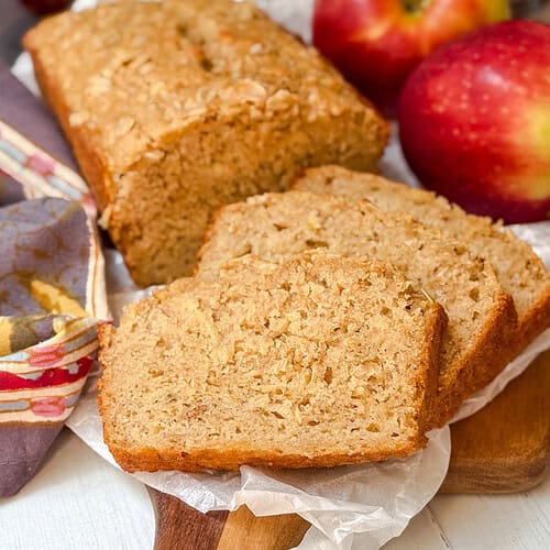 Two slices of apple oatmeal bread rest in front of a loaf on parchment paper, with whole apples and a patterned cloth nearby on a wooden surface.
