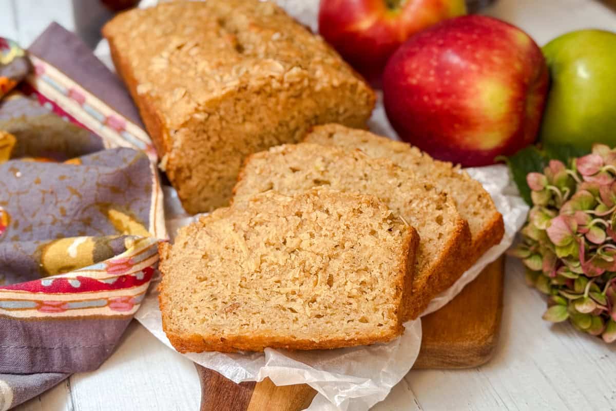 Two slices of apple oatmeal bread rest in front of a loaf on parchment paper, with whole apples and a patterned cloth nearby on a wooden surface.