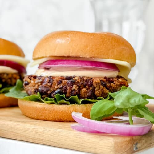 A black bean burger with lettuce, red onion, cheese, and sauce in a brioche bun sits on a wooden board, with fresh spinach leaves and onion slices beside it. Another burger and a glass are blurred in the background.