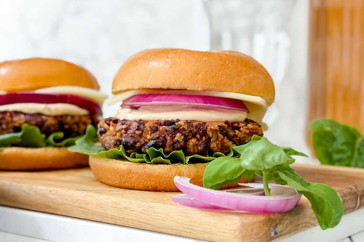 A black bean burger with lettuce, red onion, cheese, and sauce in a brioche bun sits on a wooden board, with fresh spinach leaves and onion slices beside it. Another burger and a glass are blurred in the background.
