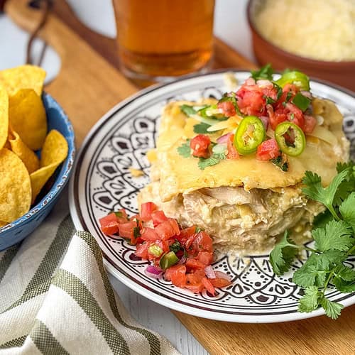 A plate with a serving of chicken enchilada casserole topped with jalapeños and pico de gallo, garnished with cilantro, next to tortilla chips, salsa, cheese, and sour cream-a perfect Crock Pot comfort meal.