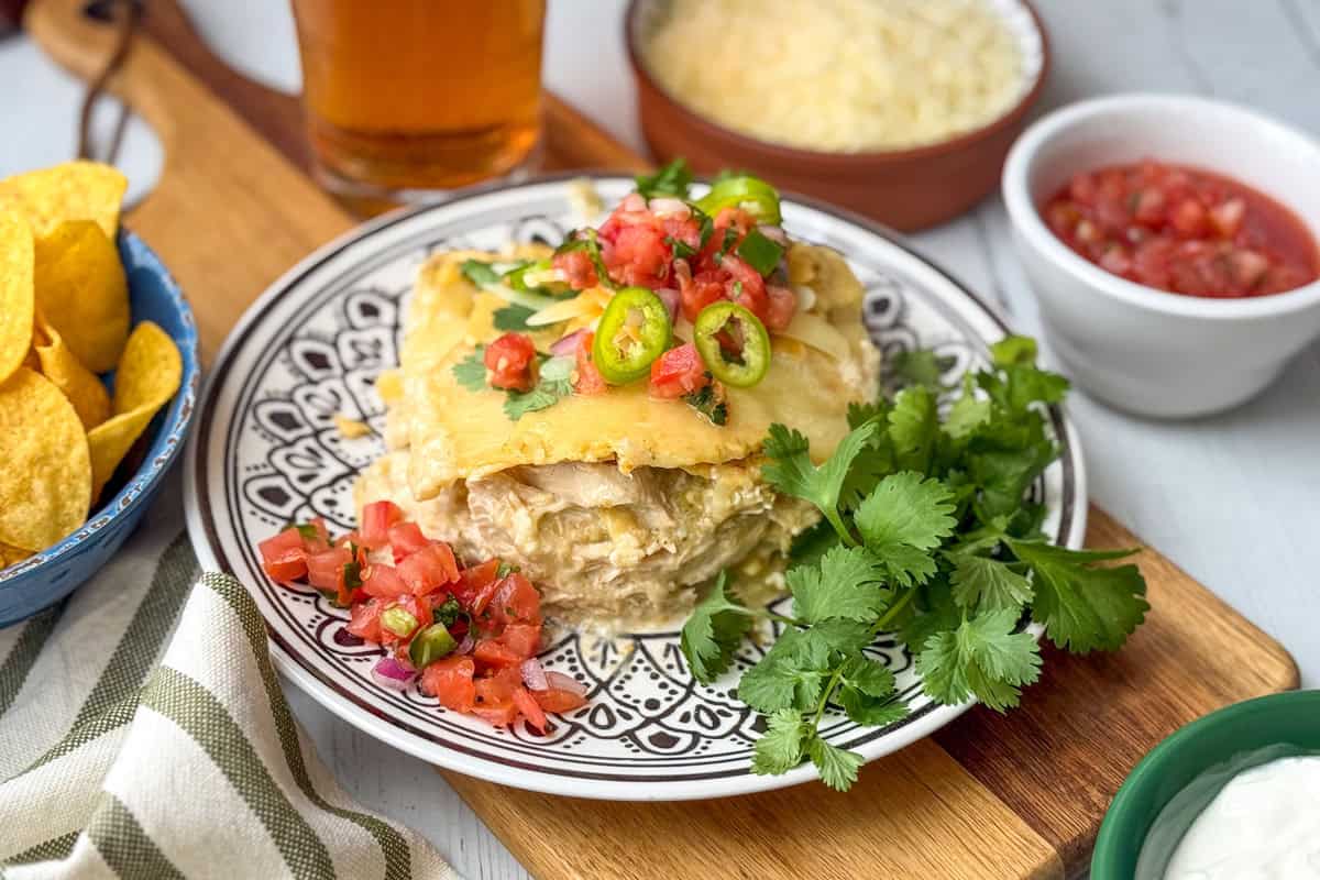 A plate with a serving of chicken enchilada casserole topped with jalapeños and pico de gallo, garnished with cilantro, next to tortilla chips, salsa, cheese, and sour cream-a perfect Crock Pot comfort meal.