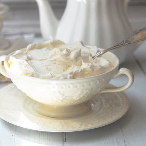 A white bowl filled with homemade clotted cream and a spoon sits on a matching saucer on a light wooden table. Stacked teacups and a teapot appear in the background.