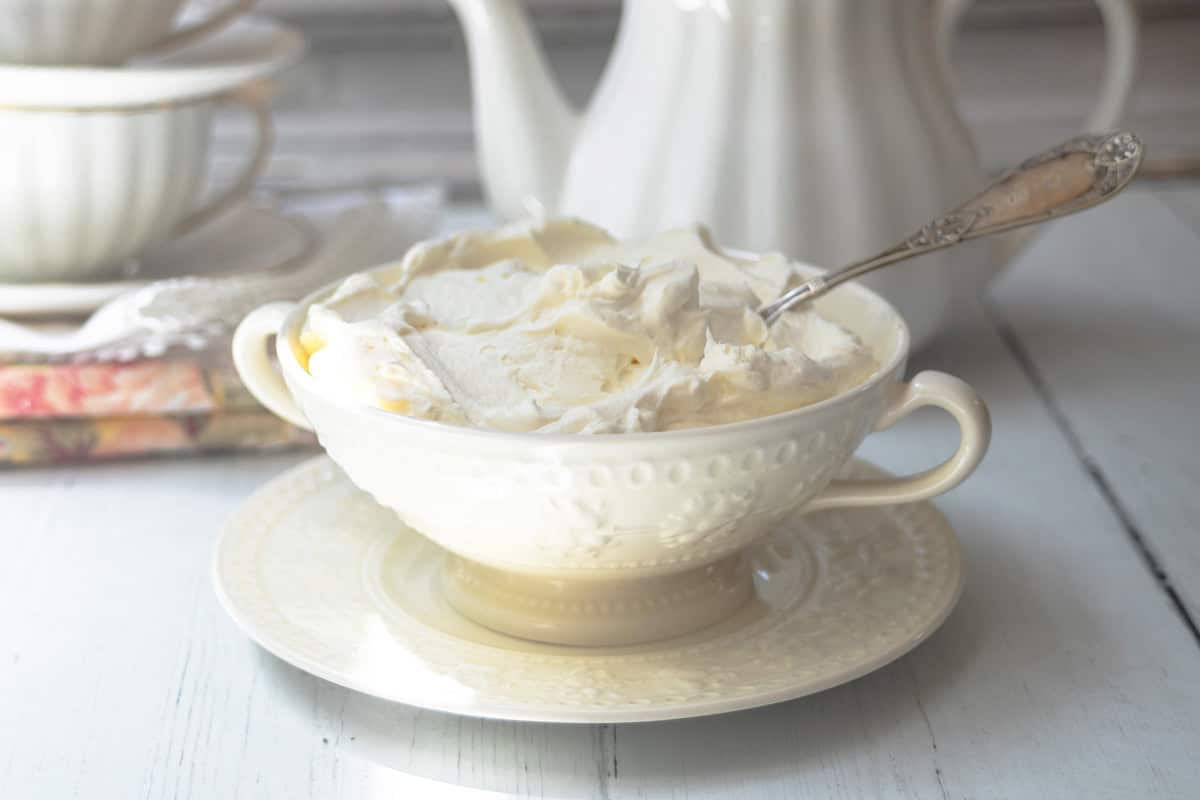 A white bowl filled with homemade clotted cream and a spoon sits on a matching saucer on a light wooden table. Stacked teacups and a teapot appear in the background.