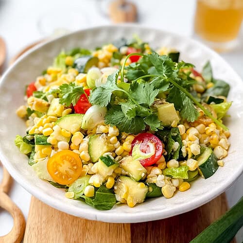 A bowl of fresh corn salad with cherry tomatoes, avocado, chopped greens, cucumber, cilantro, and hints of grilled zucchini, served on a light wooden board with utensils and a drink in the background.