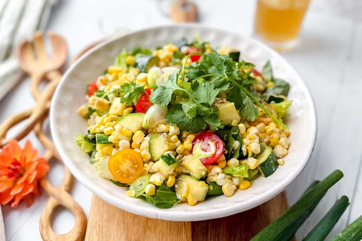 A bowl of fresh corn salad with cherry tomatoes, avocado, chopped greens, cucumber, cilantro, and hints of grilled zucchini, served on a light wooden board with utensils and a drink in the background.