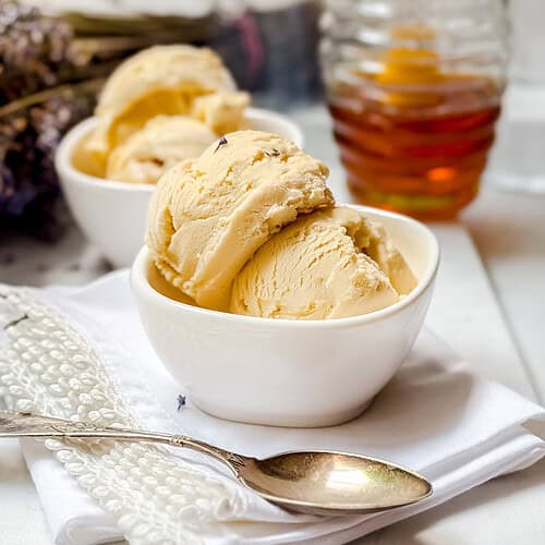 Two white bowls filled with scoops of creamy yellow ice cream sit on a folded white napkin, with a silver spoon beside them. A jar of honey and dried lavender flowers are in the background on a white surface.