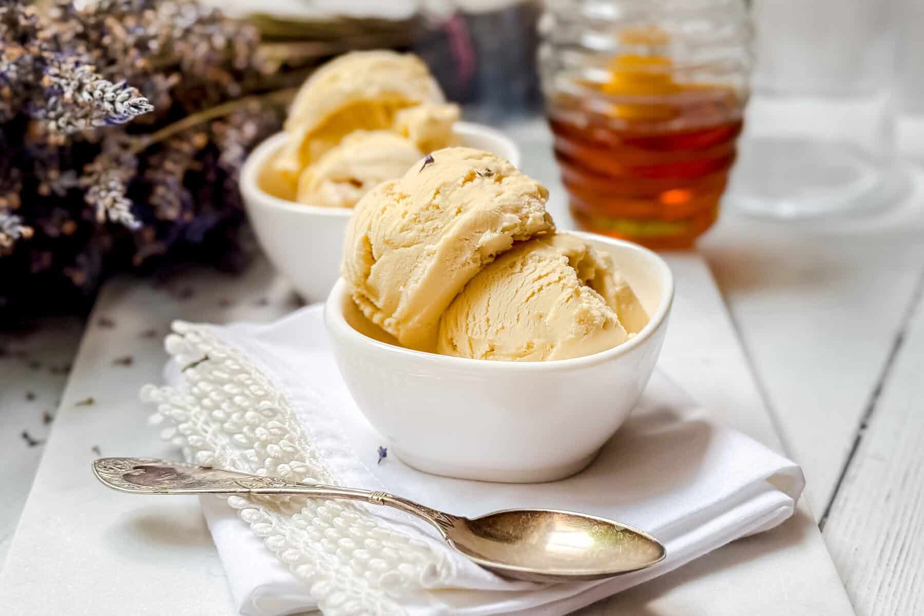 Two white bowls filled with scoops of creamy yellow ice cream sit on a folded white napkin, with a silver spoon beside them. A jar of honey and dried lavender flowers are in the background on a white surface.